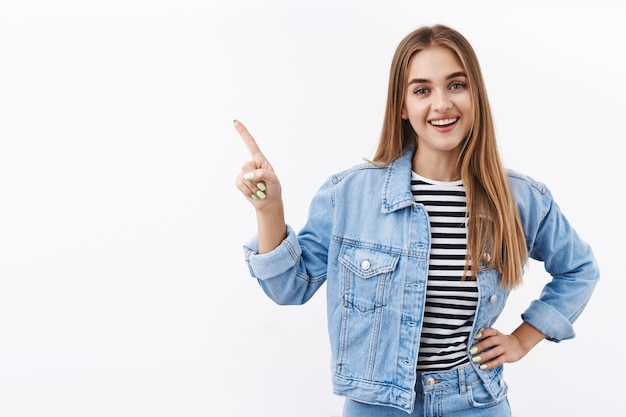 Carefree good-looking blond woman pointing upper left corner and talking to at camera with broad satisfied smile, recommend awesome place, great online store, standing on white wall joyful