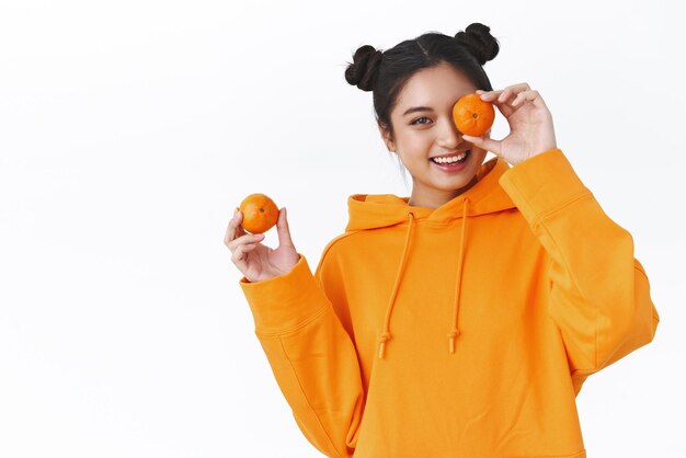 Беззаботная симпатичная молодая азиатская девушка с двумя пучками волос в оранжевой толстовке, держащая мандарины и улыбающаяся, игриво поедающая фрукты, заботясь о здоровье, применяя натуральную косметику на белом фоне