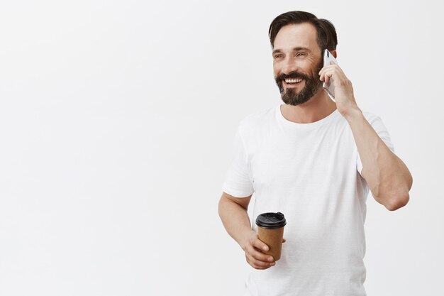 Carefree handsome adult man drinking coffee and talking on smartphone