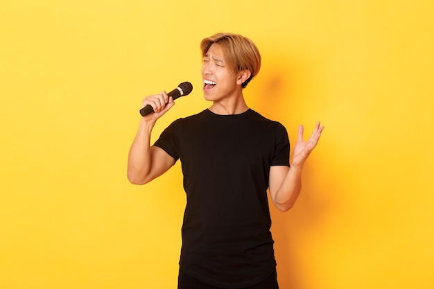 屈託のないハンサムなアジア人、韓国の歌手が黄色の壁に立って、情熱を持ってマイクに向かって歌う