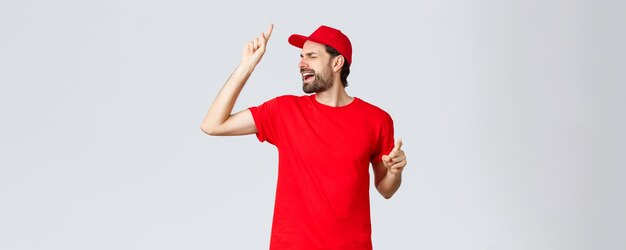 赤いTシャツとキャップでのんきなハンサムなひげを生やした男の配達の従業員は目を閉じて歌うraisi ...