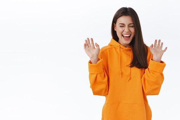 Carefree happy brunette woman in orange hoodie close eyes and scream from happiness and joy win trip to paradise island finally going to vacation raising hands up in celebration