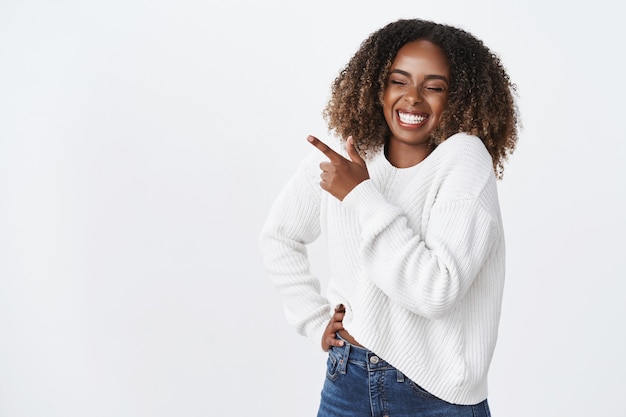 Carefree happy dark-skinned plump young woman curly hairstyle wear sweater hold hand pocket carefree feel joyful smiling laughing happily pointing upper left corner funny promo, amusing ad