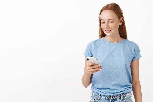 喜びとデバイスの画面を見つめる新しいスマートフォンを介して青いTシャツのテキストメッセージでそばかすのある屈託のない幸せで楽しげな魅力的な赤毛の女性