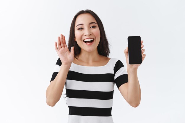 のんきな幸せで熱狂的な格好良いアジアの女性は、スマートフォンアプリケーションの空白の携帯電話のディスプレイを笑顔で喜んで見せて友好的に手を振るように片手を上げるカジュアルな縞模様のTシャツを着ています