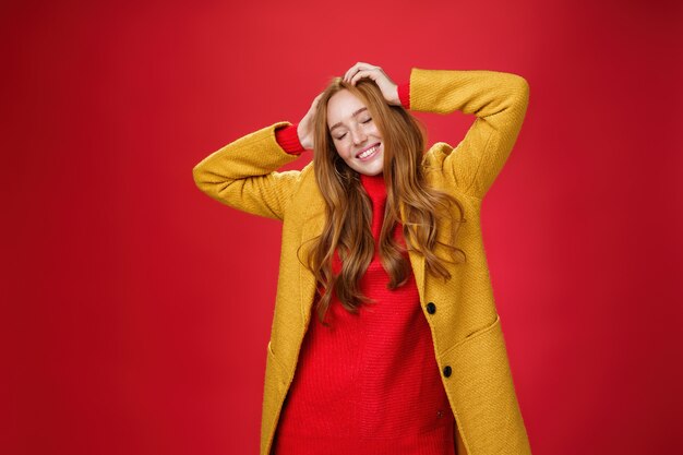 のんきな幸せな生姜の女の子は、音楽と良い雰囲気で手をつないで目を閉じて、黄色いコートの赤い背景に対して踊り、楽しんで喜んでリラックスして笑っています。