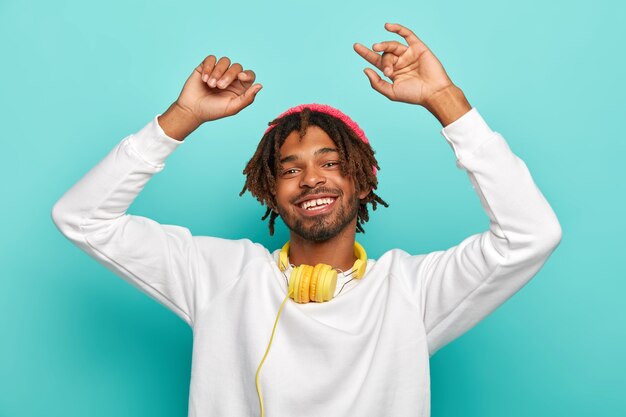 Carefree happy hipster listens music in modern headset, raises arms and dances joyfully, feels relaxed, wears white jumper, isolated over blue background