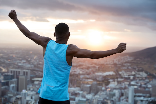 Carefree independent man with dark skin, stands back, stretches hands as holding world, feels freedom, wears blue casual vest, enjoys panoramic view with sunrise. Recreation concept