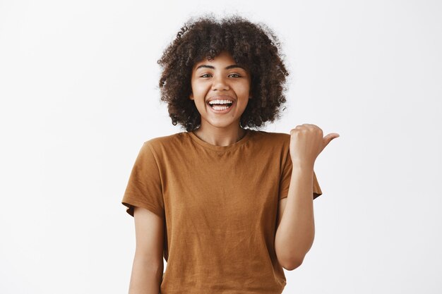 carefree joyful and attractive young dark-skinned woman in stylish t-shirt pointing right and smiling broadly while showing way or asking question about curious thing