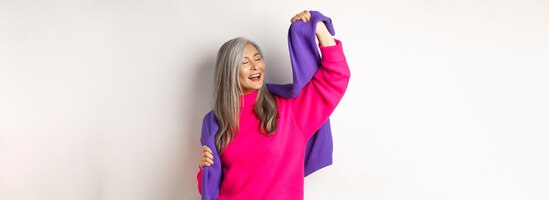 Free photo carefree korean elderly woman in pink sweater dancing with sweatshirt on shoulders and smiling posin