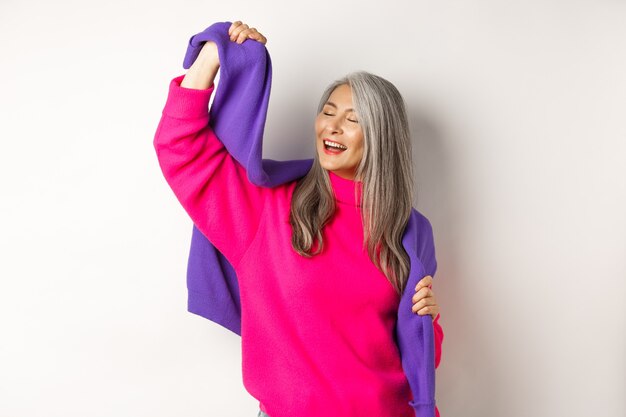 Carefree korean elderly woman in pink sweater, dancing with sweatshirt on shoulders and smiling, posing happy over white background