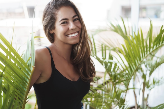 Carefree lovely tanned young woman standing balcony near green plant smiling laughing out loud look adorable grin camera talking casually coworker veranda enjoy perfect sunny summer days