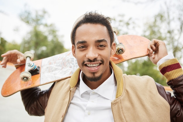 Carefree mixed race male sketaboarder holds skateboard behind, smiles happily