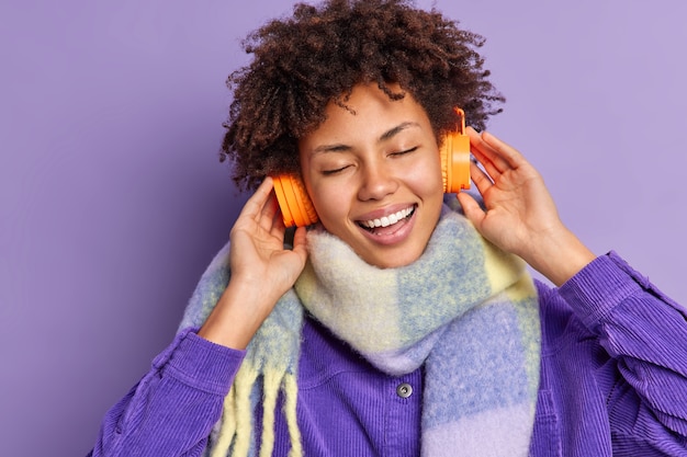 Free photo carefree optimistic afro american teenage girl smiles broadly keeps hands on stereo headphones listens music keeps eyes closed wears warm scarf around neck.