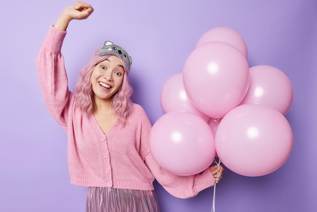 Carefree optimistic asian woman with dyed pink hair dances on party holds bunch of inflated airballoons wears sleepmask and festive clothes moves against purple background happy holidays concept