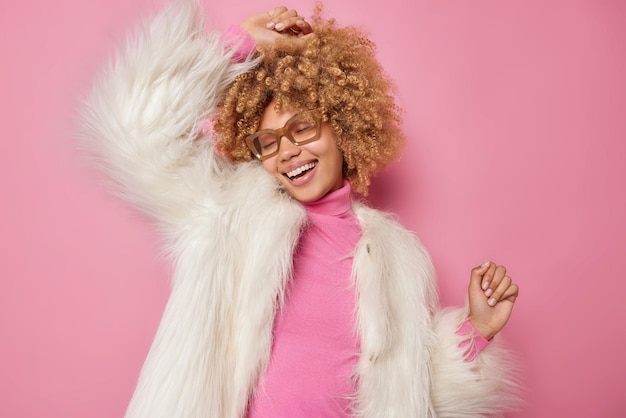Бесплатное фото Беззаботная оптимистичная привлекательная женщина с вьющимися волосами трясет руками, танцует под музыку, носит повседневную водолазку и белую шубу, радостно улыбается, движется к розовой стене студии, полная счастья