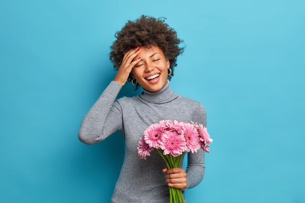 Carefree optimistic dark skinned ethnic woman holds bouquet of beautiful flowers smiles broadly