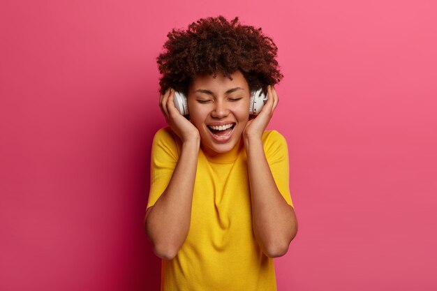 Carefree optimistic young woman smiles broadly, keeps eyes closed, shows white teeth, listens audio track, wears headphones on ears, enjoys every bit of new favourite song, laughs positively