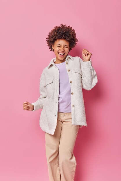 Carefree overjoyed woman dances and laughs happily has upbeat mood dressed in fashionable clothes isolated over pink wall