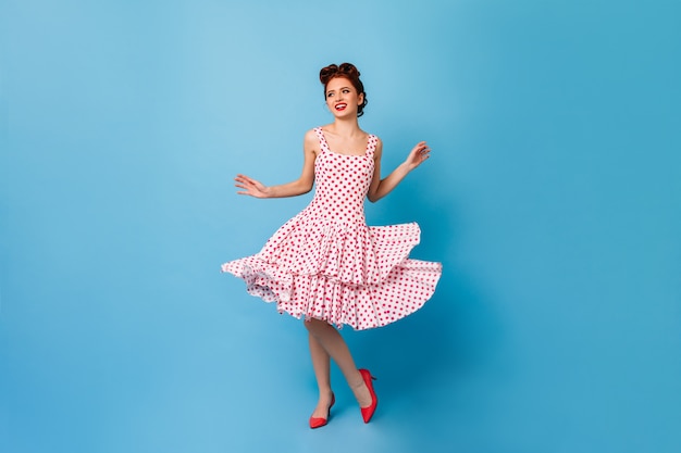 Carefree pinup girl dancing and laughing. Cute young woman in polka-dot dress having fun on blue space.