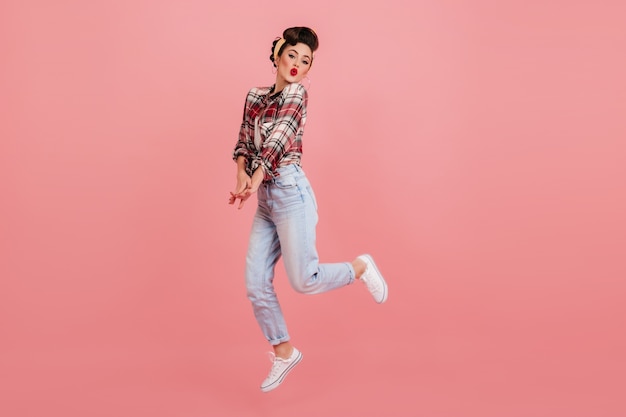 ピンクの背景にジャンプするのんきなピンナップガール。ジーンズと市松模様のシャツを着た素敵な若い女性のスタジオショット。