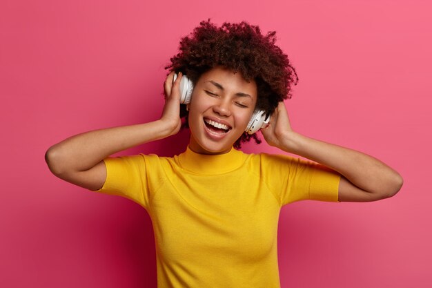 Carefree pleased millennial girl listens song in headphones, enjoys playlist, moves with rhythm, smiles broadly, keeps eyes shut, isolated on pink wall. People, leisure, lifestyle, hobby concept