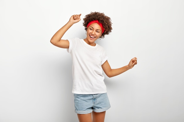 Carefree pleased teenage girl has fun, dances joyfully with raised arms, being entertained and amused, wears summer clothes