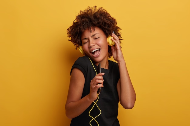 Carefree positive curly woman hears awesome track via headset, sings aloud with song, holds smartphone as microphone