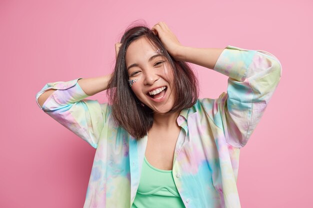 Carefree positive pretty Asian teenage girl keeps hands on head moves actively enjoys life smiles broadly wears casual clothes feels amused has fun during spare time isolated over pink wall