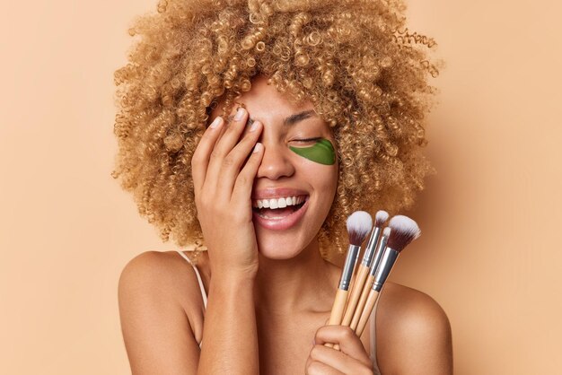 のんきなポジティブな若い女性は、顔の手のひらを喜んで笑わせ、目を閉じて広く笑顔を保ちます。ベージュの壁に隔離された化粧ブラシで化粧をする緑のヒドロゲルパッチを適用します
