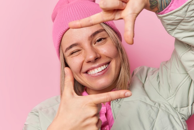 Carefree positive young woman makes frame gesture finds perfect angle winks eye and smiles broadly wears jacket and hat poses against pink background looks through fingers measures something