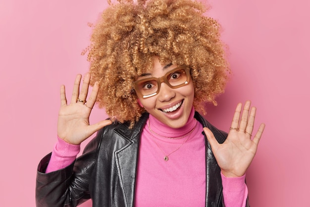 Бесплатное фото Беззаботная позитивная молодая женщина с вьющимися густыми волосами держит ладони поднятыми вперед, улыбается камера, счастливо выражает счастливые эмоции, носит очки и повседневную одежду, изолированную на розовом фоне.