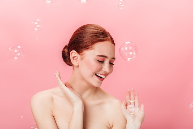 비누 거품과 함께 포즈 평온한 예쁜 소녀입니다. 분홍색 배경에 웃 고 매력적인 생강 여자의 스튜디오 샷.