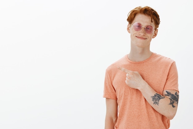 Carefree redhead man in sunglasses smiling, pointing upper left corner pleased