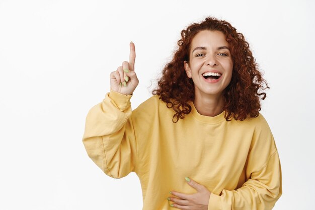 Carefree redhead woman laughing, pointing finger up at smth funny, smiling happy at camera, white background