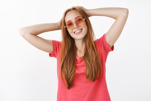 トレンディなサングラスと頭の後ろに手を繋いでいるピンクのtシャツを着てのんきなリラックスして幸せな若い赤毛の女性