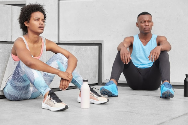 Carefree restful black sister and brother deep in thoughts, have break after jogging, wear casual comfortable clothes, surrounded with bottle filled with water to have energy. Youth and sport concept