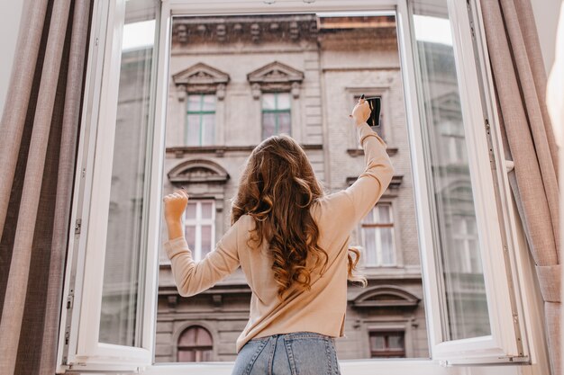 Carefree slim girl in jeans dancing beside window with cup of coffee