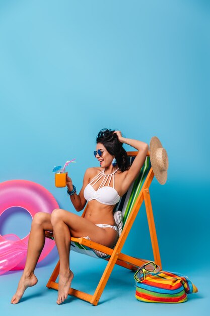 Carefree slim woman sitting on deck chair on blue background