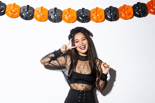 Беззаботная улыбающаяся азиатская женщина в костюме ведьмы наслаждается вечеринкой в честь Хэллоуина, танцует и показывает жест мира, стоя на белом фоне с украшением из тыквы.