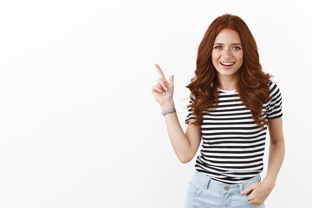 Carefree smiling cute redhead woman in striped tshirt smiling delighted discuss cool new offer pointing upper left corner indicate copy space promo grin satisfied standing white background