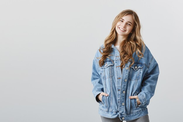 Беззаботная улыбающаяся счастливая белокурая девушка в джинсовой куртке над серой стеной