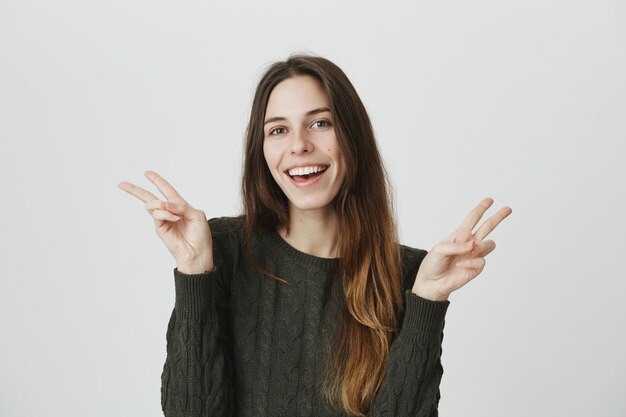 Беззаботная улыбающаяся женщина в свитере выражает позитивность, показывает знаки мира