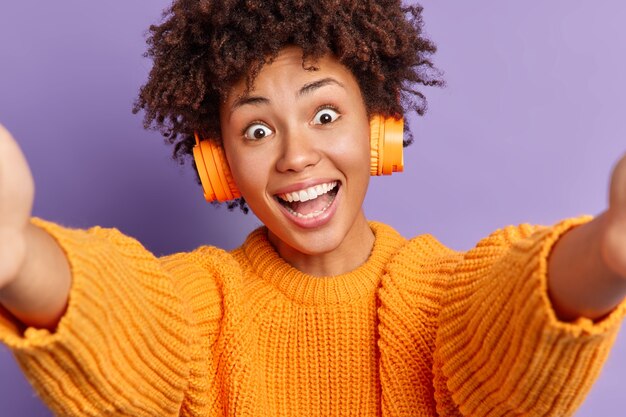 Carefree surprised happy curly African American woman looks with widely opened eyes stretches hands and takes selfie dressed in casual clothes listens favorite music via stereo wireless headphones.