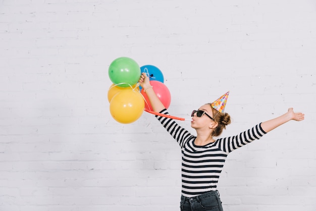 Carefree teenage girl holding colorful balloons blowing part horn standing against wall