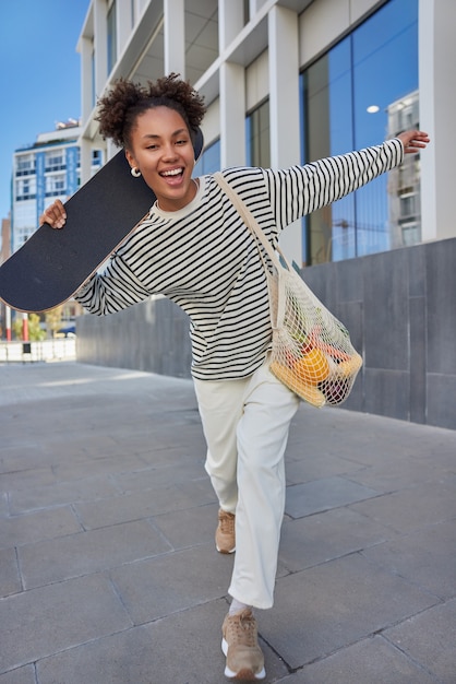 Adolescente spensierata passeggia felicemente in città tiene skateboard porta borsa a rete vestita con maglione a righe pantaloni bianchi e scarpe da ginnastica sorride felice si diverte durante la calda giornata di sole va allo skate park