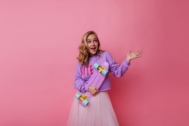 Carefree white lady in skirt holding cute longboard. Emotional female model with blonde curly hair standing on pastel.