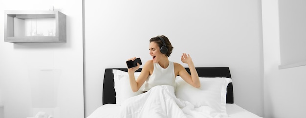 Free photo carefree woman in bed wakes up and listens to music in wireless headphones dancing and singing using
