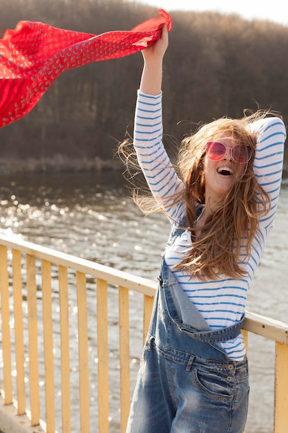 Carefree woman with sunglasses holding scarf in the wind by the lake