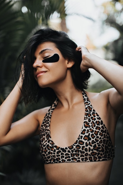 Carefree woman with tanned skin enjoying vacation. Outdoor shot of charming woman with eye patches on nature background.